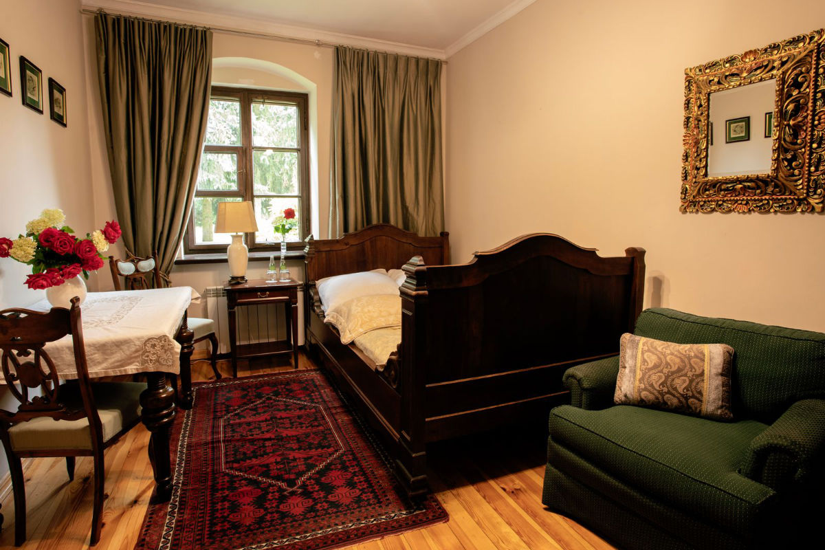 Un acogedor dormitorio con una cama individual, un sillón verde, una mesa de madera, un arreglo floral y una alfombra. Una ventana con cortinas verdes deja pasar la luz natural y un espejo decorativo cuelga de la pared.