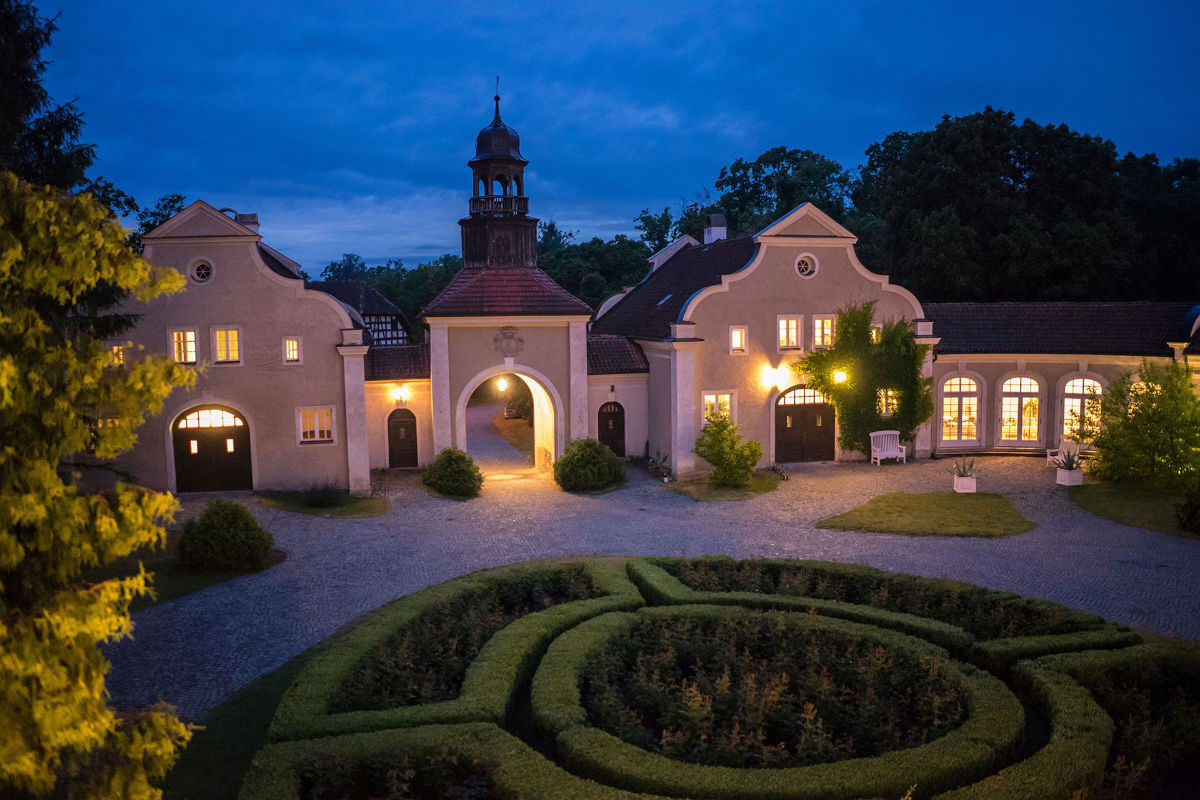 The evening view of the elegant Palac i Folwark Galiny showcases its central archway, glowing windows, and a beautifully manicured circular garden in the foreground.
