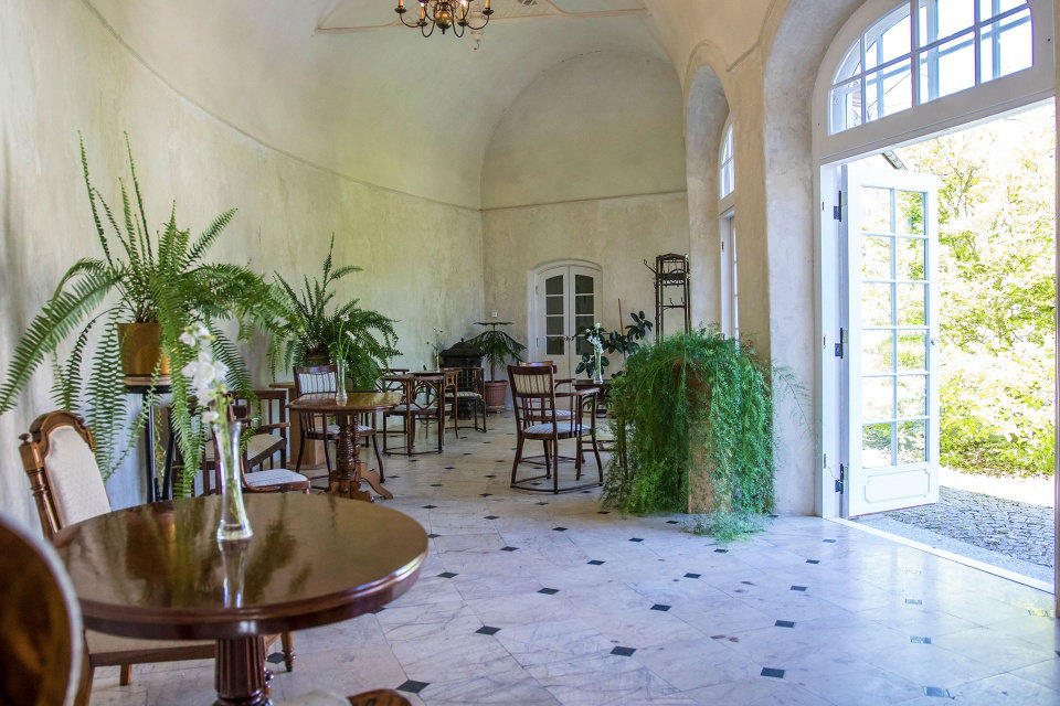 Una sala illuminata dal sole con piante in vaso, tavoli e sedie in legno su un pavimento piastrellato evoca il fascino del Palac i Folwark Galiny. Grandi finestre e porte si aprono su una tranquilla vista sul giardino.