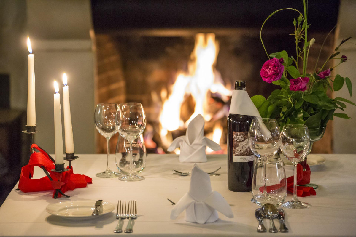 Un tavolo da pranzo apparecchiato per due persone con bicchieri da vino Palac i Folwark Galiny, tovaglioli piegati e una bottiglia di vino si trova accanto a un vaso di fiori. Candele accese e un accogliente caminetto completano l'ambiente intimo sullo sfondo.