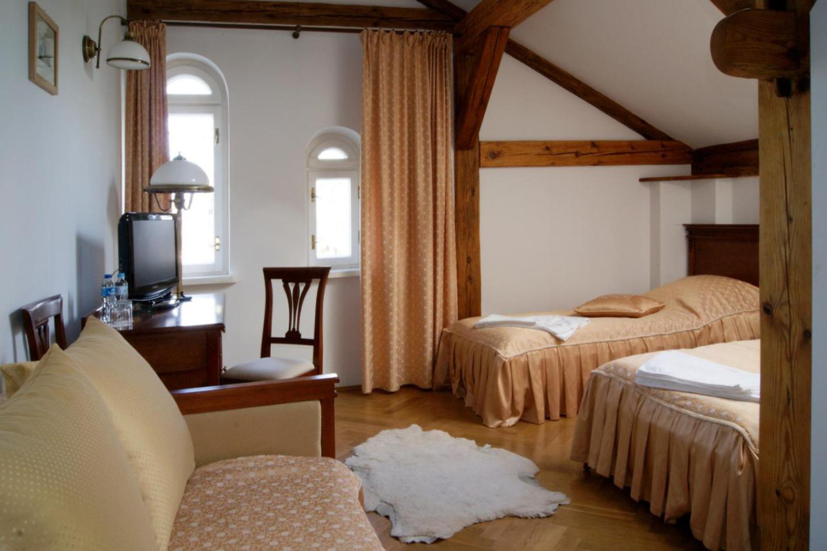 Accogliente camera del Pałac i Folwark Łochów con due letti singoli, un piccolo divano, travi in legno e tende color pesca sulle finestre ad arco. La TV si trova sulla scrivania in legno, mentre un tappeto bianco abbellisce il pavimento in legno.