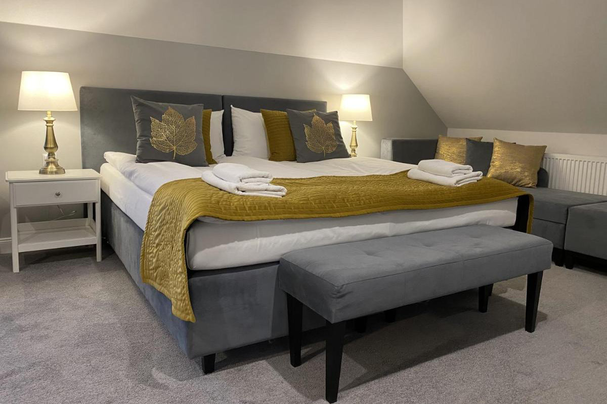 Un letto ben fatto con accenti dorati, due comodini con lampade e asciugamani piegati sopra evocano l'eleganza di Pałac i Folwark Łochów. Una panca grigia si trova ai piedi del letto.