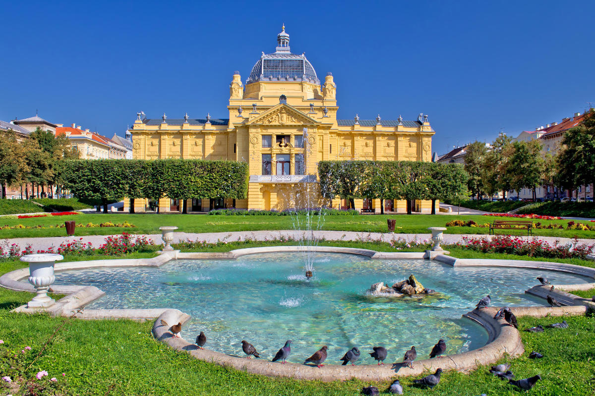 An ornate yellow art pavilion with a domed roof stands majestically behind a fountain in Zagreb, Croatia, surrounded by a garden brimming with flowers and pigeons under the bright, clear sky.