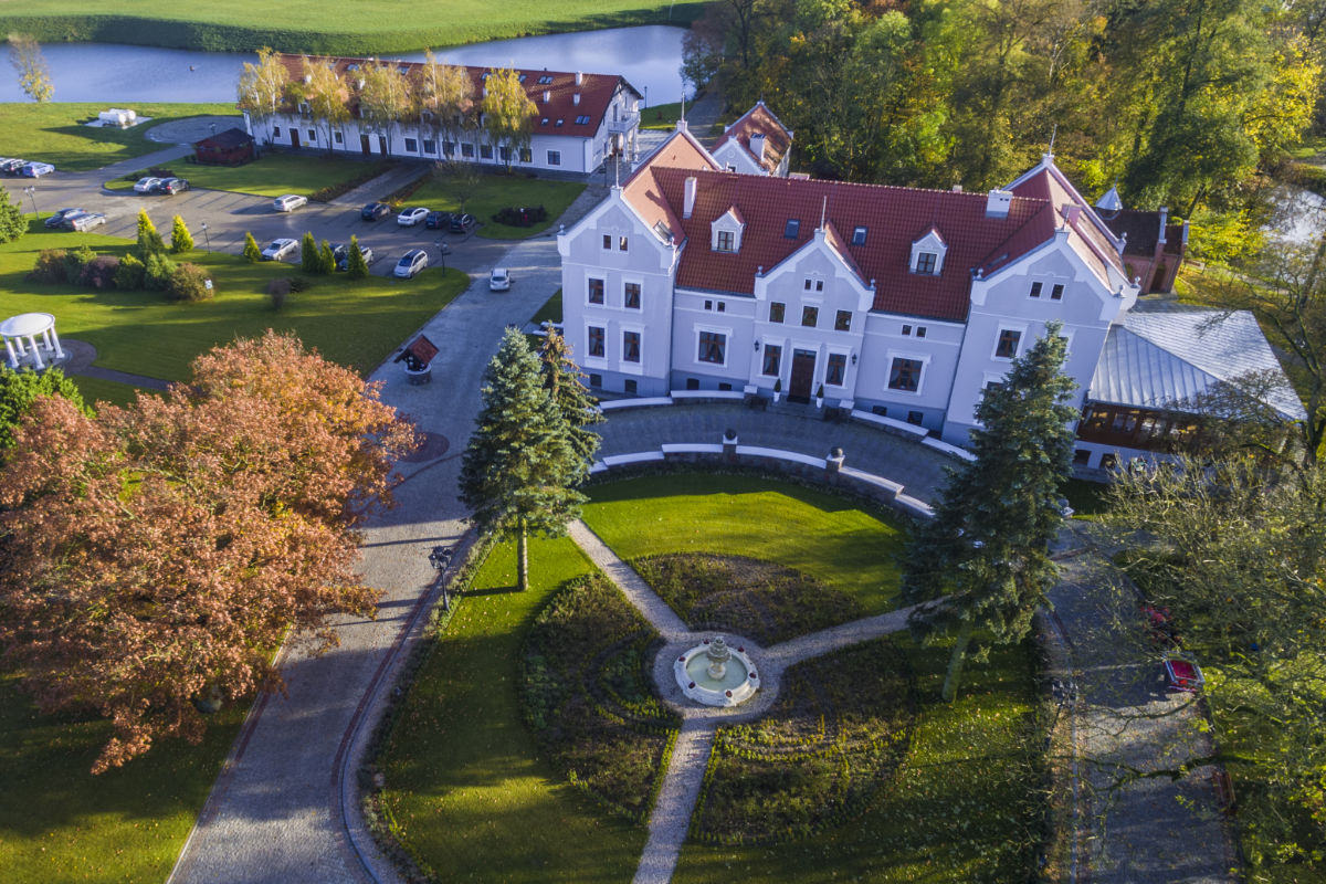 Luftaufnahme des Hotels Pałac Mortęgi, eines großen weißen Gebäudes mit rotem Dach. Es liegt inmitten von Bäumen und verfügt über eine kreisförmige Auffahrt mit einem zentralen Springbrunnen in der Nähe eines kleinen, ruhigen Sees.