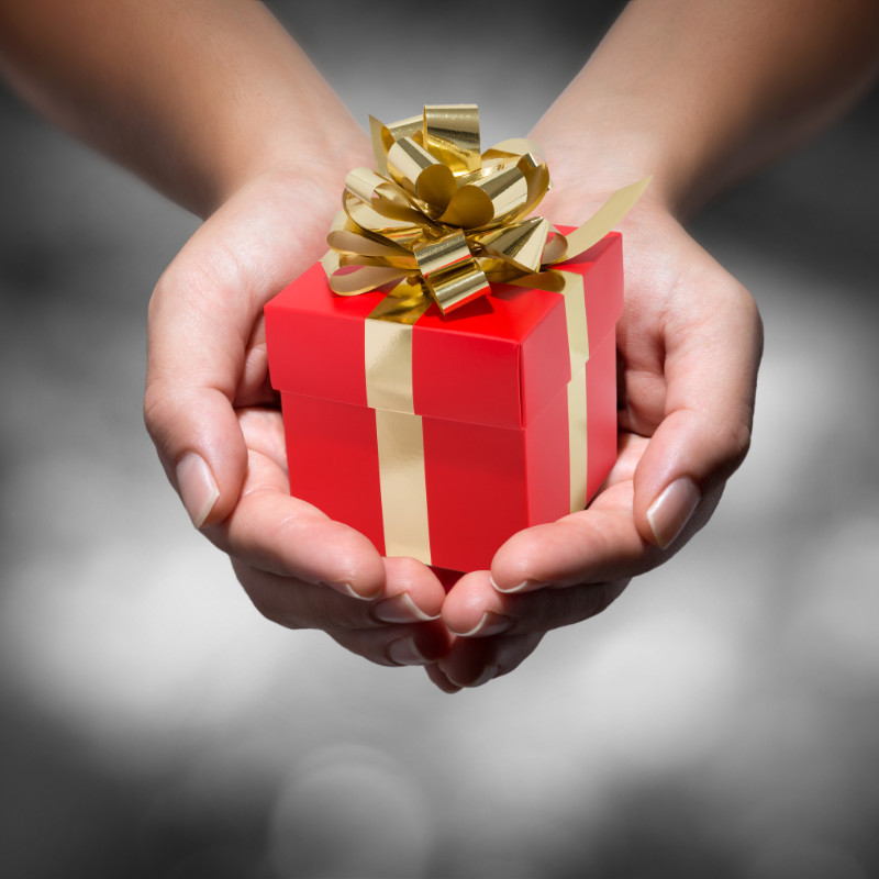 Mani che tengono in mano una piccola confezione regalo rossa con nastro e fiocco d'oro, che ricorda un caro buono regalo, su uno sfondo grigio sfocato.