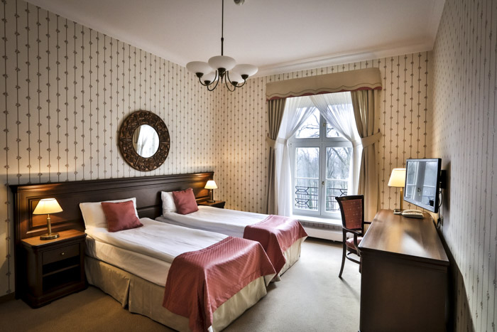 Une chambre d'hôtel avec deux lits jumeaux, des couvre-lits roses, un miroir rond, une commode en bois et une télévision. Une fenêtre avec des rideaux donne sur un balcon.