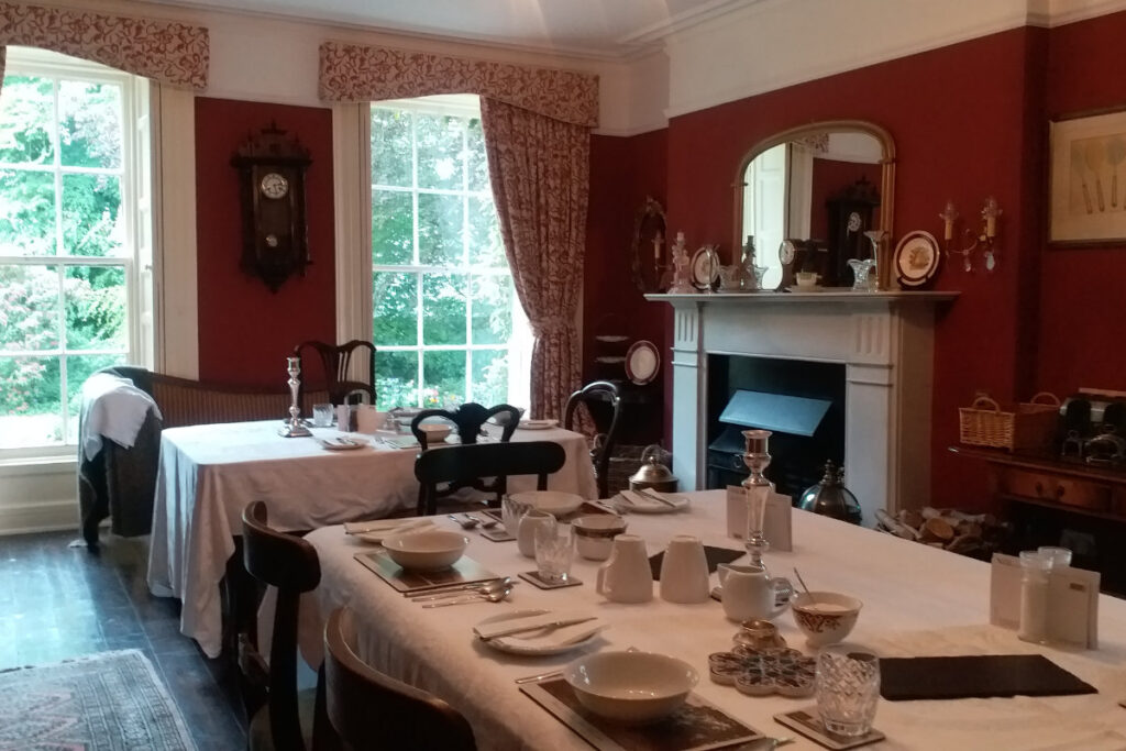 Fughe fresche: Dove alloggiare quando fa troppo caldo in città 12 Nella sala da pranzo vintage della Dromore House, le pareti rosse fanno da complemento a un accogliente caminetto e a grandi finestre. I tavoli apparecchiati con tovaglie bianche sono impreziositi da piatti e argenteria di pregio, mentre orologi e decorazioni antiche adornano le pareti e la mensola del camino.