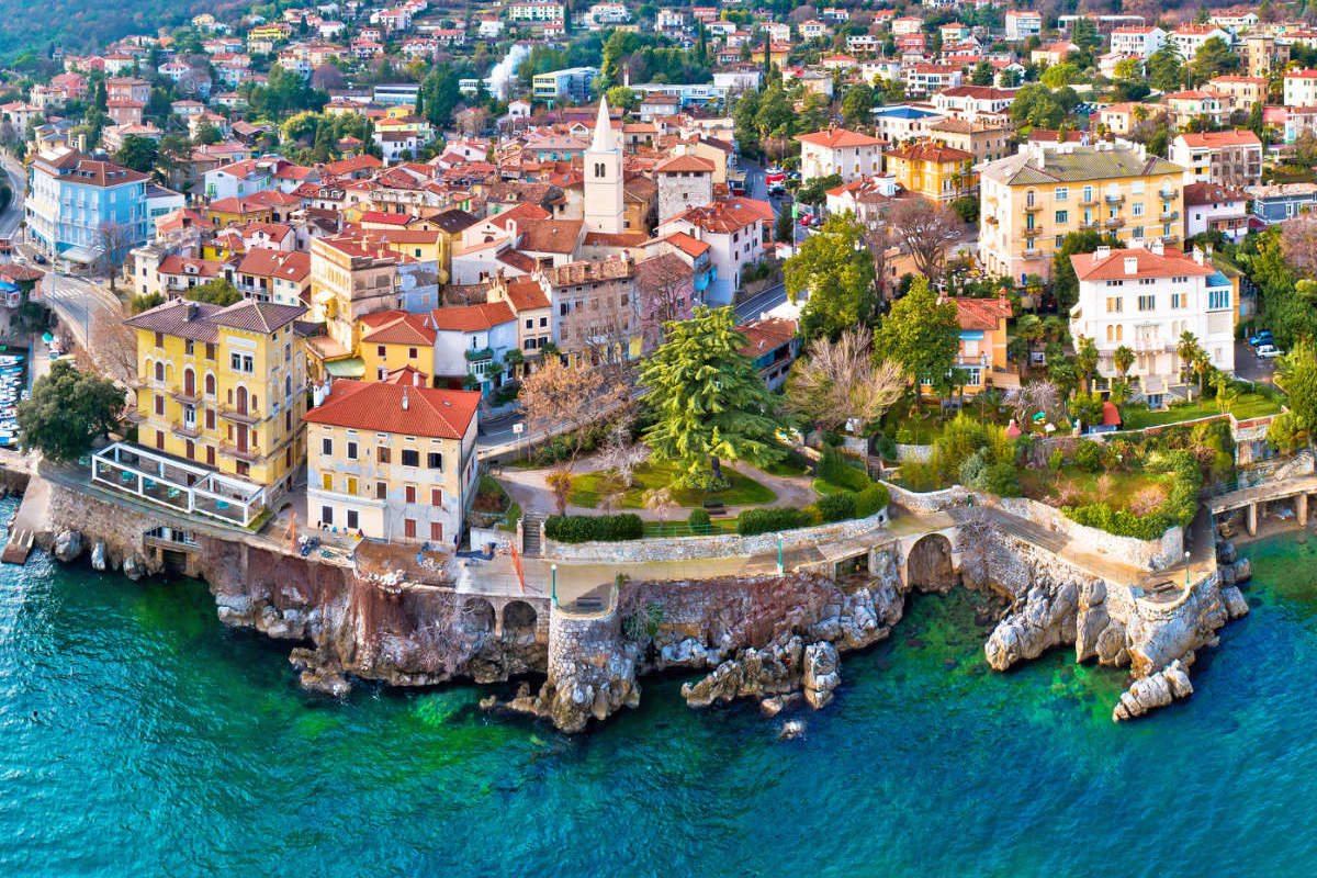 Vista aérea de Lovran (Croacia), una ciudad costera con edificios de colores, una torre de iglesia y una costa rocosa junto a aguas cristalinas de color azul verdoso.