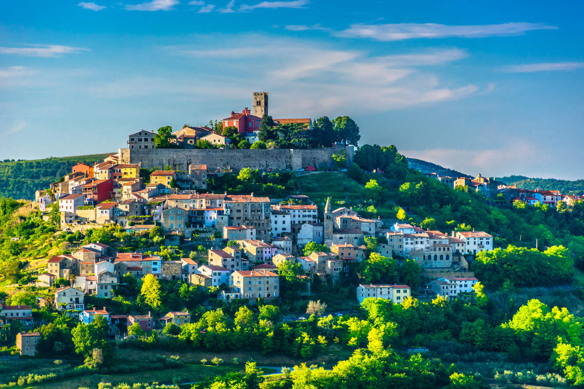Ciudad de Motovun, Croacia, en lo alto de una colina, con edificios de colores y una fortaleza de piedra, rodeada de árboles verdes bajo un cielo azul.