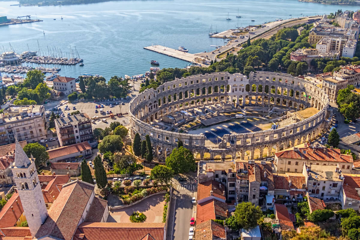 Vista aérea del antiguo anfiteatro romano de Pula, Croacia, rodeado de edificios de la ciudad, con un puerto deportivo y el paseo marítimo visibles al fondo.