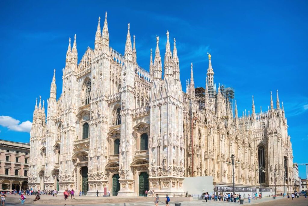 Guida di viaggio di Milano: Esplora il meglio di Milano con le nostre intuizioni locali 3 Questa cattedrale in stile gotico, descritta nella Guida turistica di Milano, vanta guglie e statue intricate. I visitatori passeggiano nella piazza antistante, sotto un cielo blu brillante.
