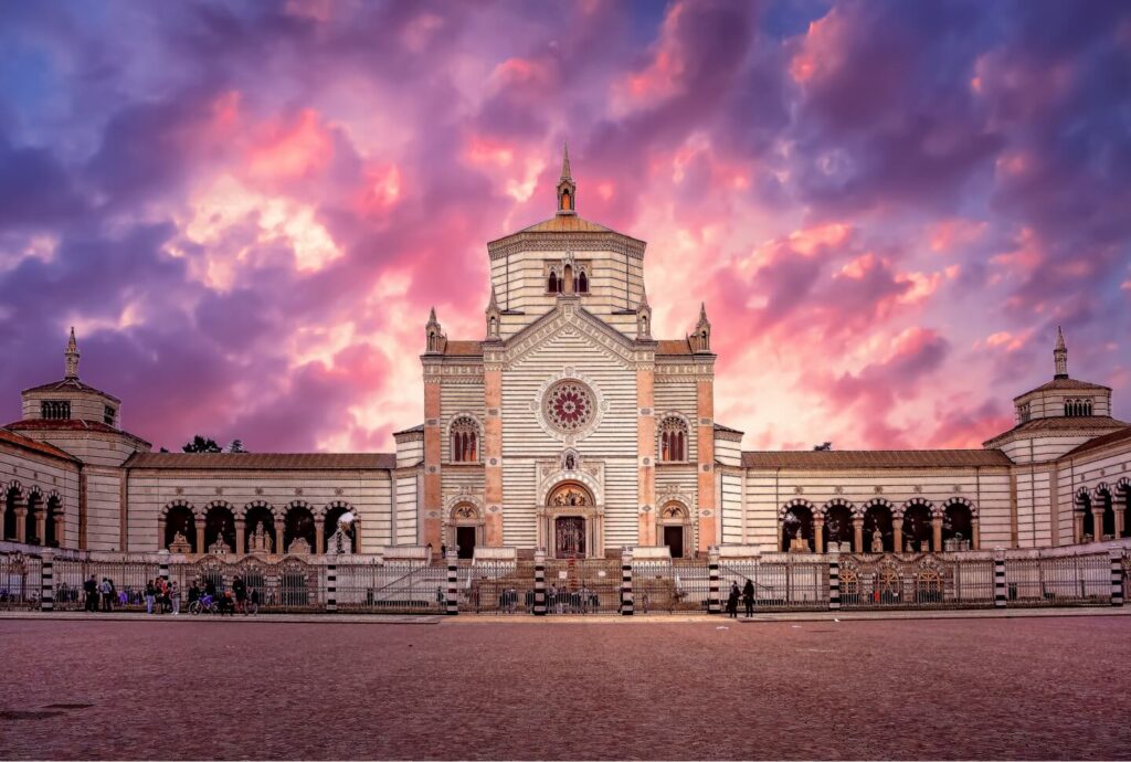 Guida di viaggio di Milano: Esplora il meglio di Milano con le nostre intuizioni locali 7 L'immagine cattura un grande edificio a strisce con dettagli ornamentali sotto un cielo vibrante e nuvoloso al tramonto, illustrando una tappa obbligata in qualsiasi guida turistica di Milano. Gli archi e la torre centrale della struttura si ergono maestosi, mentre persone e alberi animano lo sfondo.