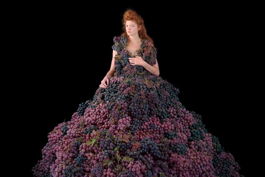 Une femme est assise, vêtue d'une robe volumineuse entièrement composée de raisins violets et rouges, sur un fond noir uni.