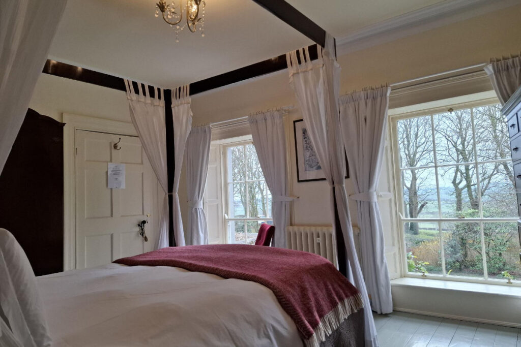 Fughe fresche: Dove alloggiare quando fa troppo caldo in città 11 L'accogliente camera da letto della Dromore House è dotata di un letto a baldacchino ornato da tende bianche e da una coperta rossa, mentre le ampie finestre offrono un'incantevole vista su un giardino alberato.