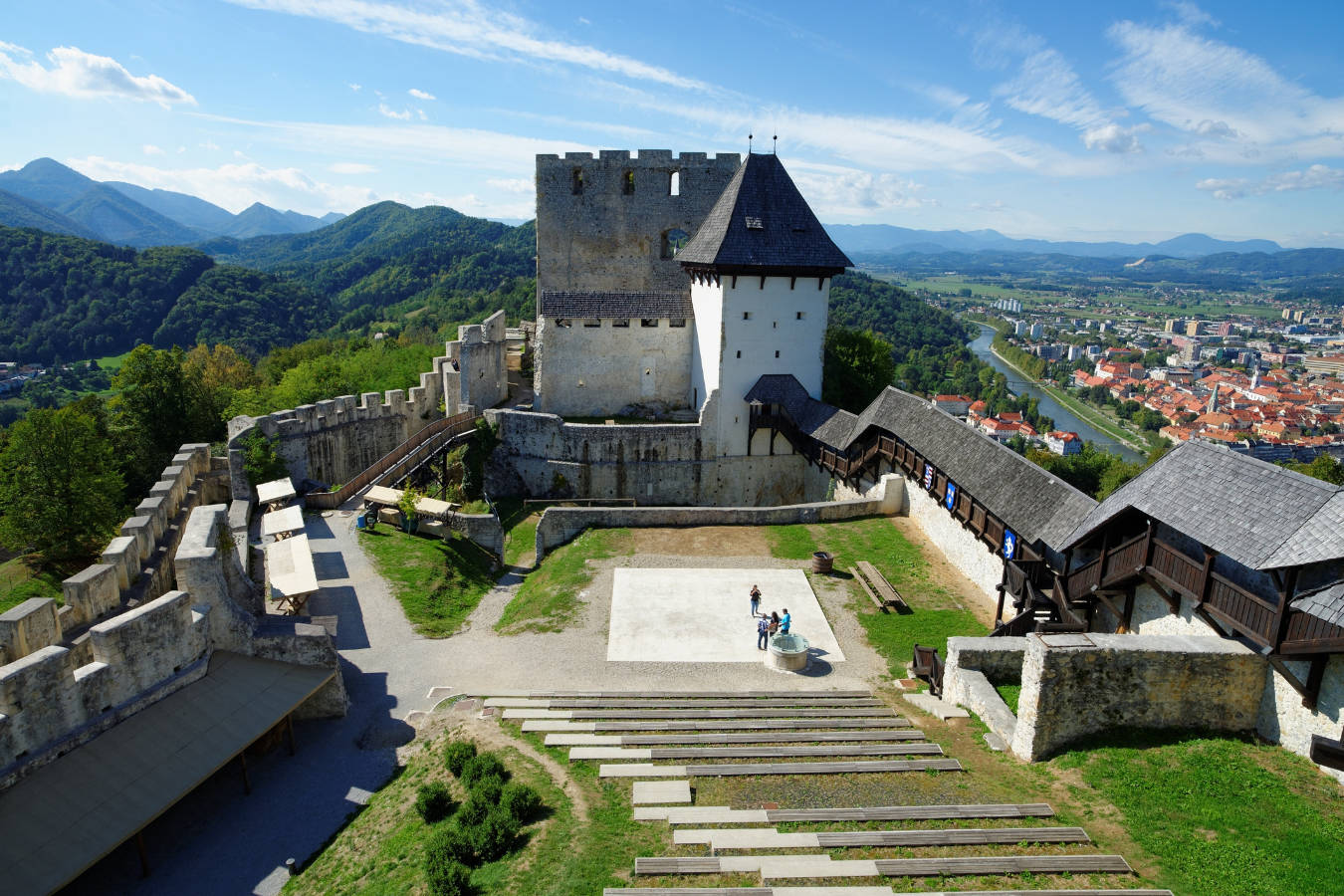 Le château de Celje, en Slovénie, une grande forteresse en pierre avec une tour centrale, se trouve au sommet d'une colline entourée de montagnes verdoyantes et d'une ville en arrière-plan. Quelques personnes se tiennent dans la cour ouverte.