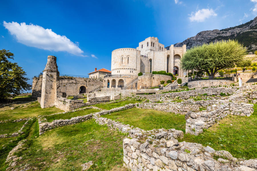 Kruja Castle, with its round and rectangular towers, stands amid ruins and lush greenery, set against distant mountains beneath a blue sky with scattered clouds.