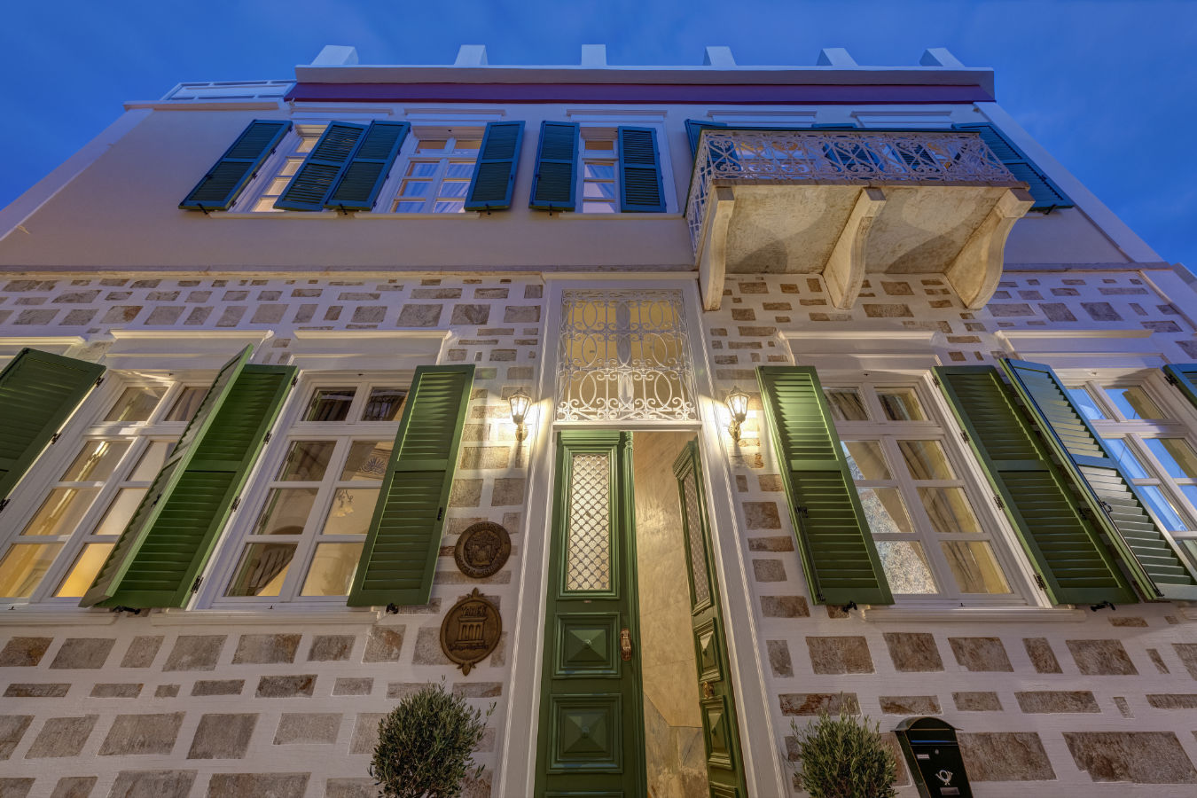 Vue extérieure du Palazzo Santa Maria, un bâtiment en pierre de deux étages avec des volets verts, une porte verte, un balcon et un éclairage chaleureux, photographié au crépuscule.