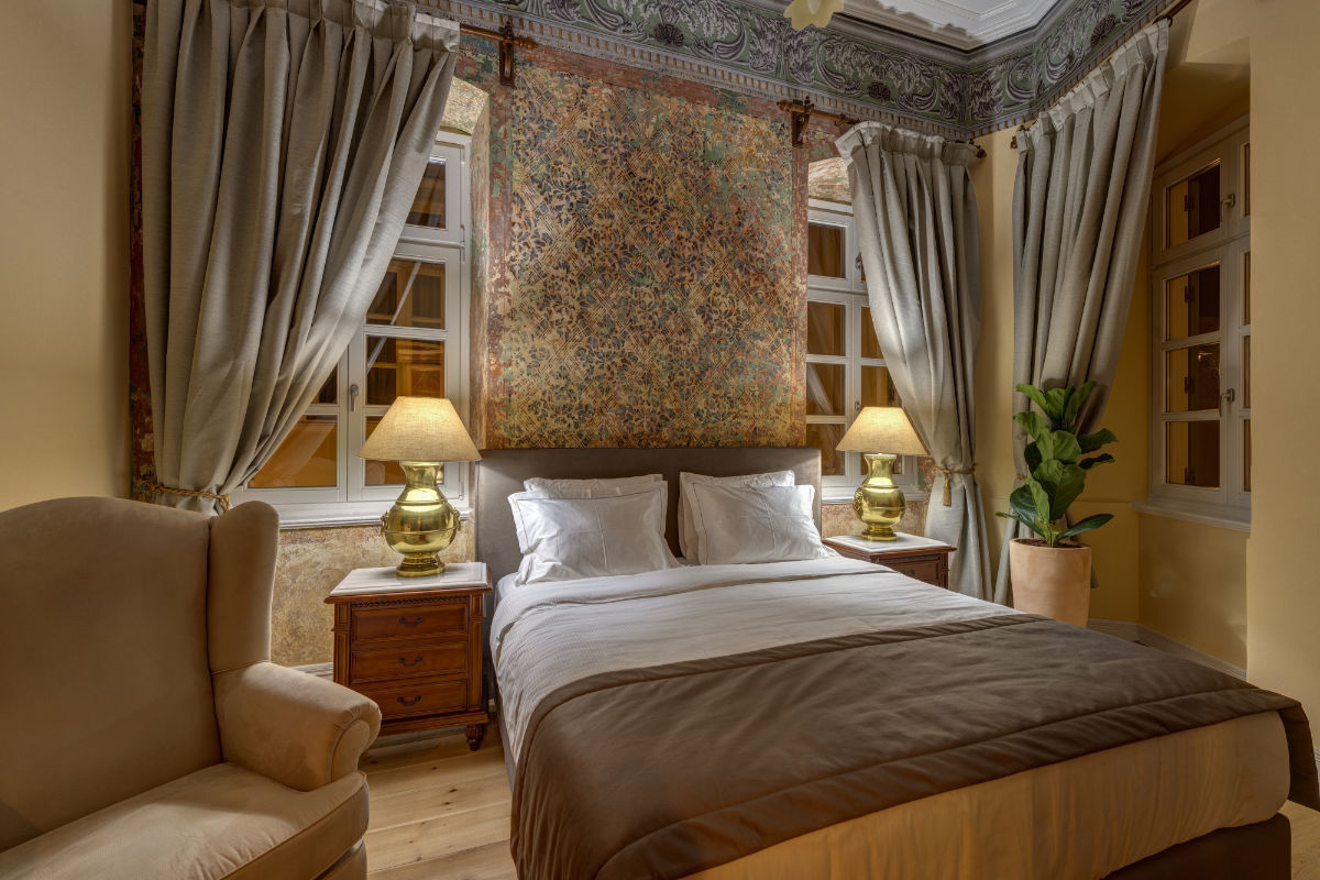 Una acogedora habitación del Palazzo Santa Maria cuenta con una cama de matrimonio, dos mesillas de noche con lámparas, un sillón, ventanas con cortinas, una maceta y adornos decorativos cerca del techo.