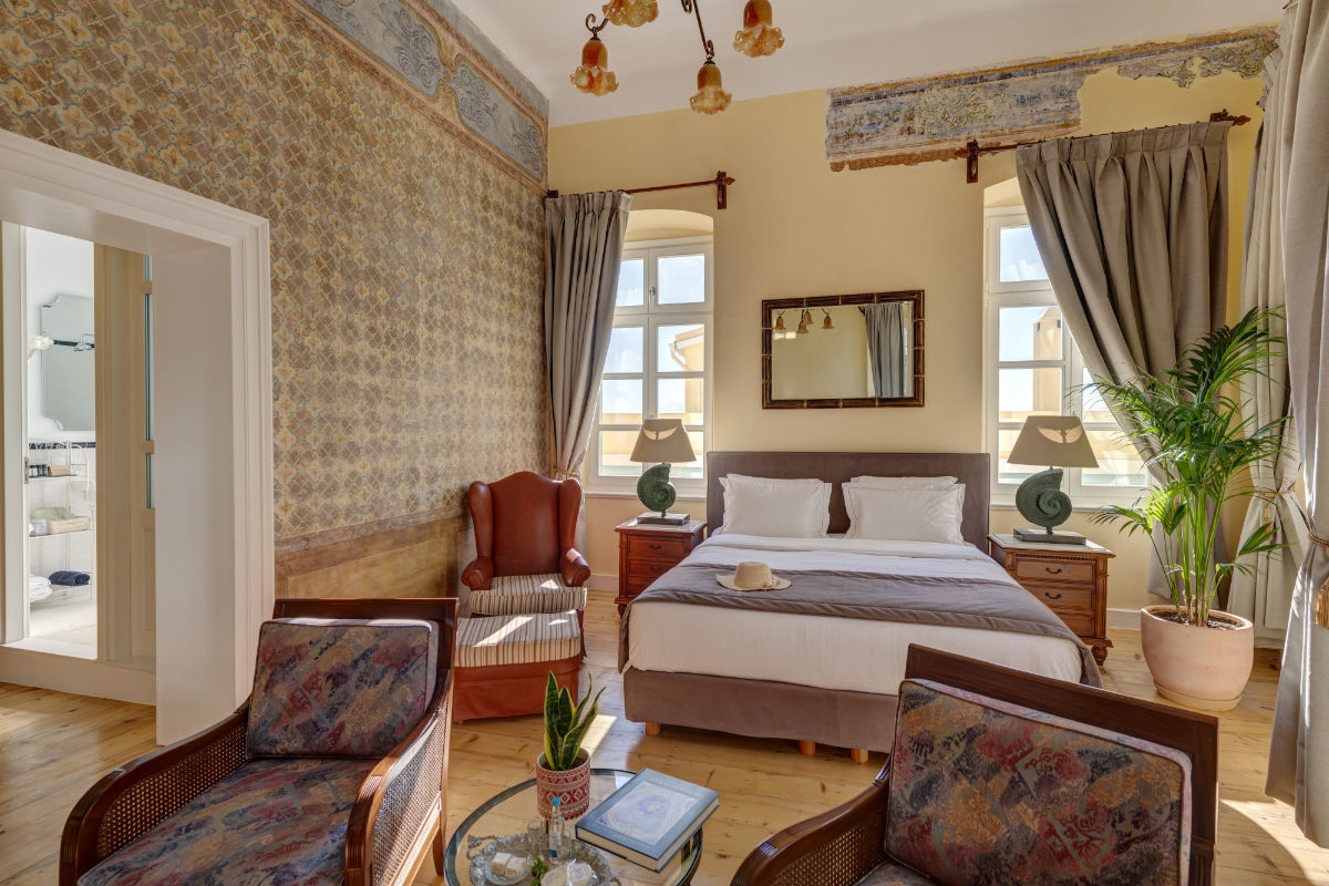 Un dormitorio bien iluminado del Palazzo Santa Maria cuenta con papel pintado estampado, una cama doble, dos sillones, una silla de cuero con respaldo, una maceta y dos ventanas con cortinas. A través de una puerta abierta se ve un cuarto de baño.