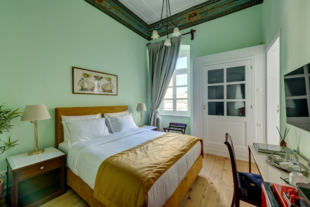 Una cama bien hecha con detalles amarillos en un dormitorio verde menta del Palazzo Santa Maria, con ventana, escritorio, mesilla de noche y molduras decorativas en el techo.