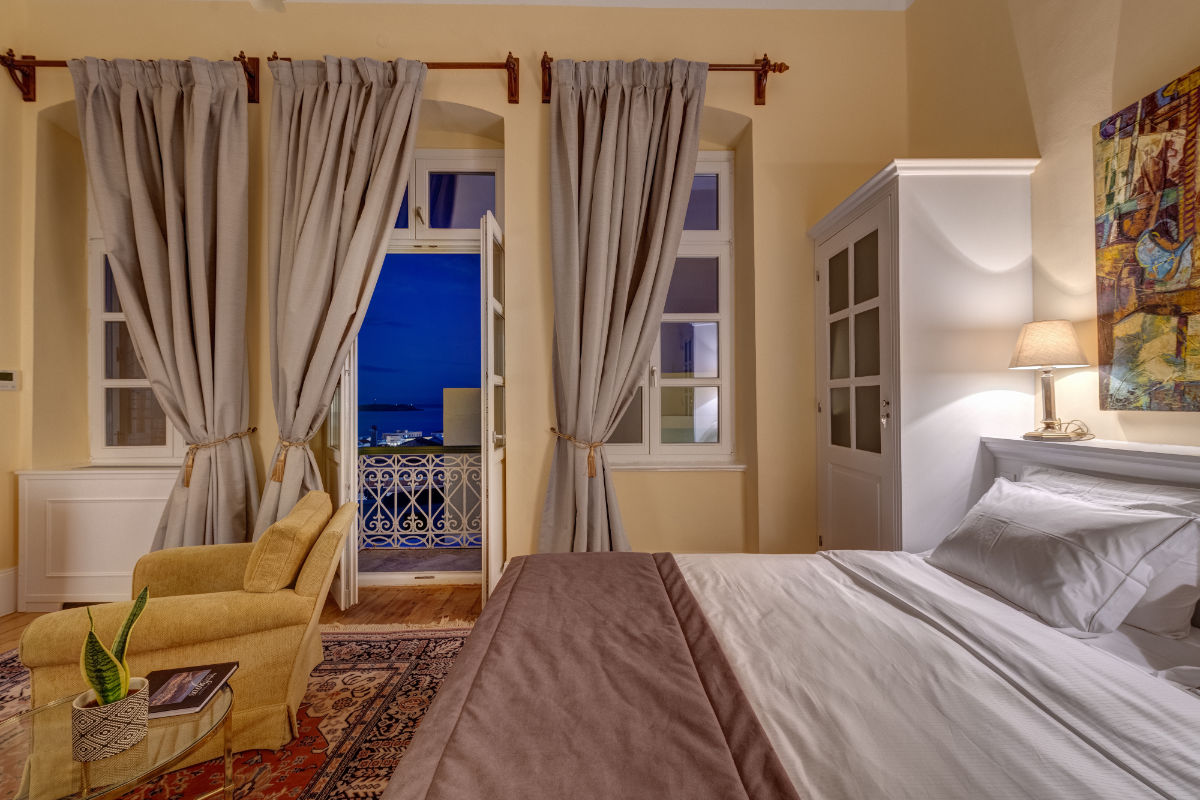 Elegante dormitorio en el Palazzo Santa Maria con una cama grande, sillón, planta en maceta y puerta abierta a un balcón con vistas nocturnas. Los tonos neutros y la iluminación tenue crean un ambiente acogedor.