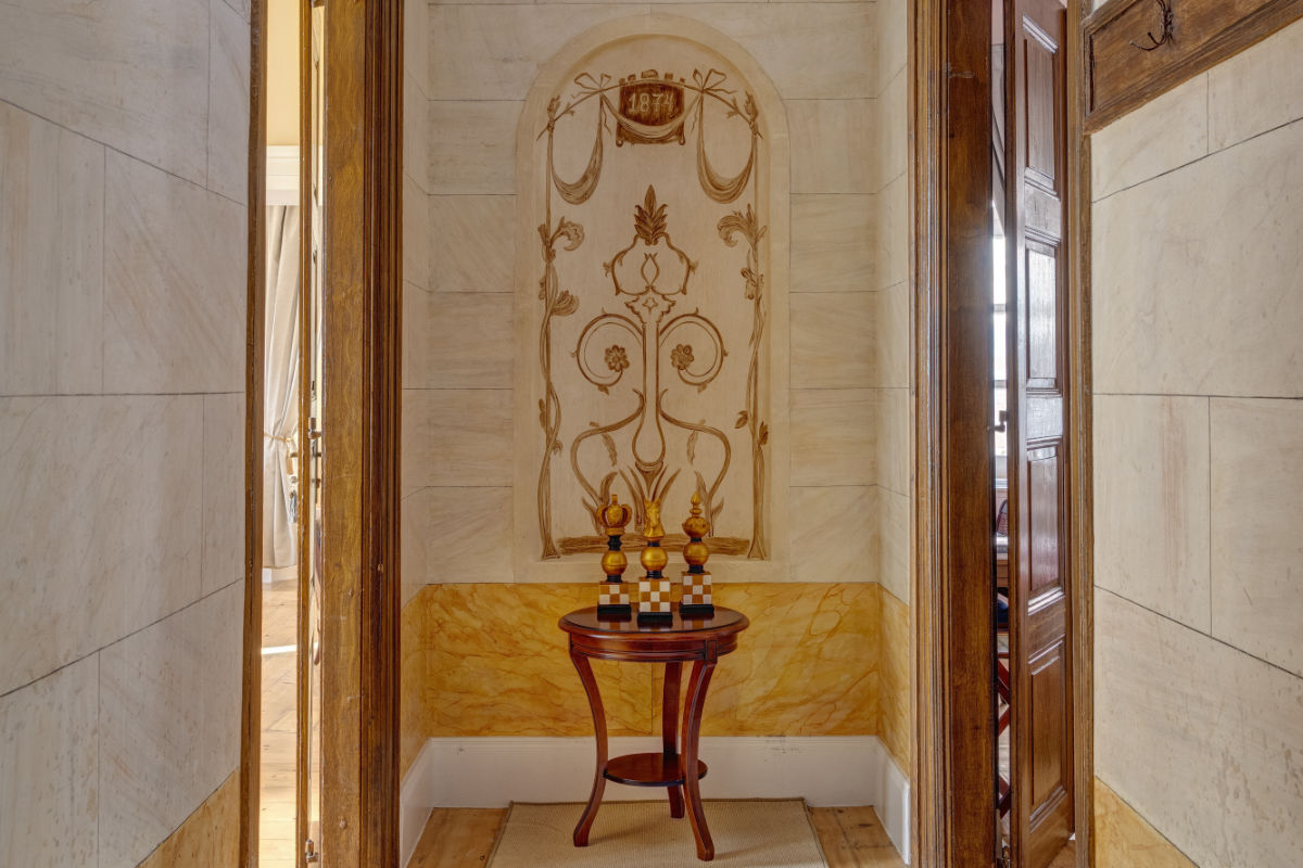 Una pequeña mesa de madera con piezas de ajedrez decorativas se encuentra en un estrecho pasillo con paredes decoradas con tallas y paneles de madera que evocan la refinada elegancia del Palacio Santa María.