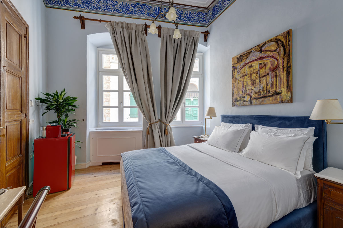 Una cama bien hecha con ropa de cama azul y blanca en un luminoso dormitorio del Palazzo Santa Maria, con grandes ventanales, paredes beige, una planta en maceta y un cuadro abstracto sobre el cabecero.