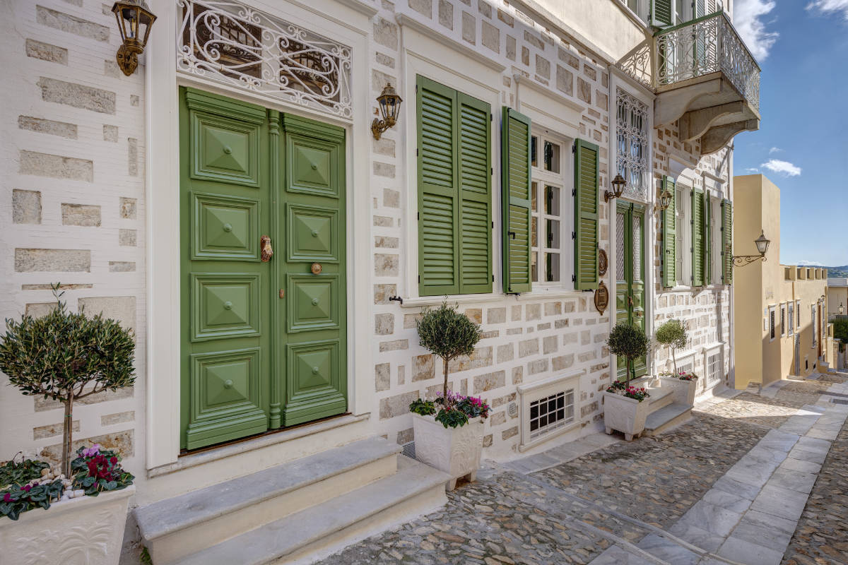 Ein traditionelles Steinhaus, das an den Palazzo Santa Maria erinnert, mit grünen Türen und Fensterläden, Topfpflanzen am Eingang und einer Kopfsteinpflasterstraße unter freiem Himmel.