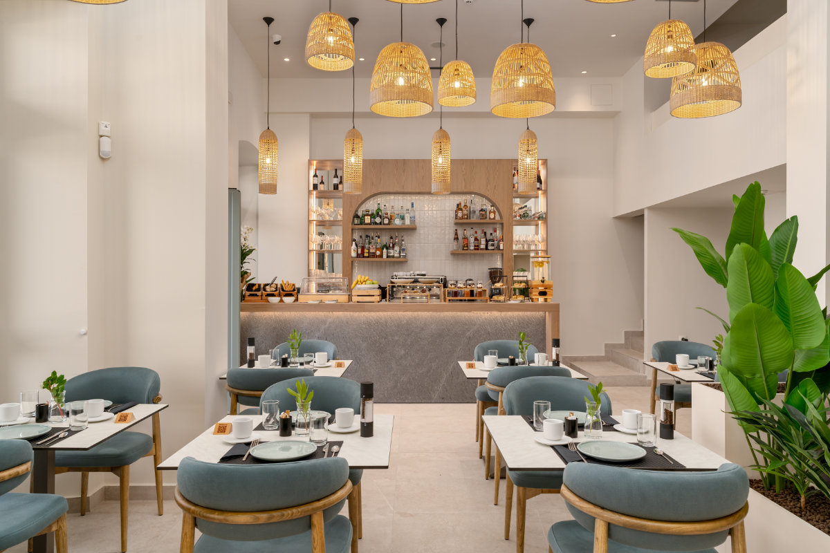 Un restaurant moderne et bien éclairé au YeeOn Boutique Hotel à Gythio, en Grèce, avec des chaises à coussins bleus, des tables bien rangées, des lampes en osier suspendues et un bar élégant à l'arrière-plan.