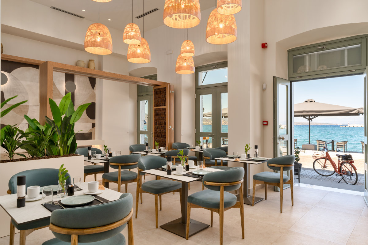 Intérieur d'un restaurant moderne avec des chaises bleues, des tables en bois, des lampes en osier suspendues et de grandes fenêtres donnant sur un patio extérieur et la mer au YeeOn Boutique Hotel ; un vélo est garé à l'extérieur dans cet hôtel de Gythio, en Grèce.