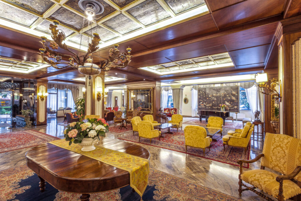 Elegant hotel lobby at the 5 star hotel Abano Terme, featuring an ornate ceiling, chandelier, antique wooden furniture, yellow upholstered chairs, floral arrangements, large rugs, and ample natural light from tall windows.
