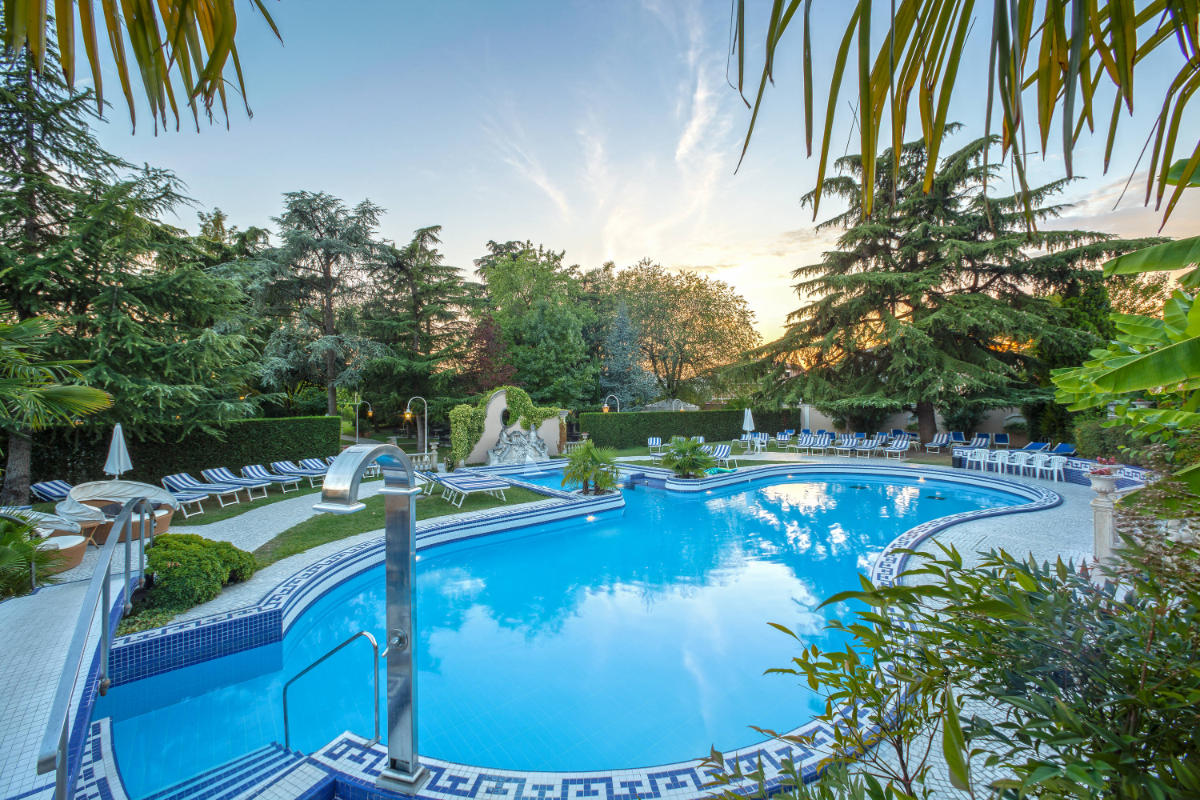 Piscine extérieure entourée de chaises longues, de parasols et de verdure luxuriante au coucher du soleil à l'Abano Ritz Hotel Terme, un hôtel 5 étoiles renommé d'Abano Terme.