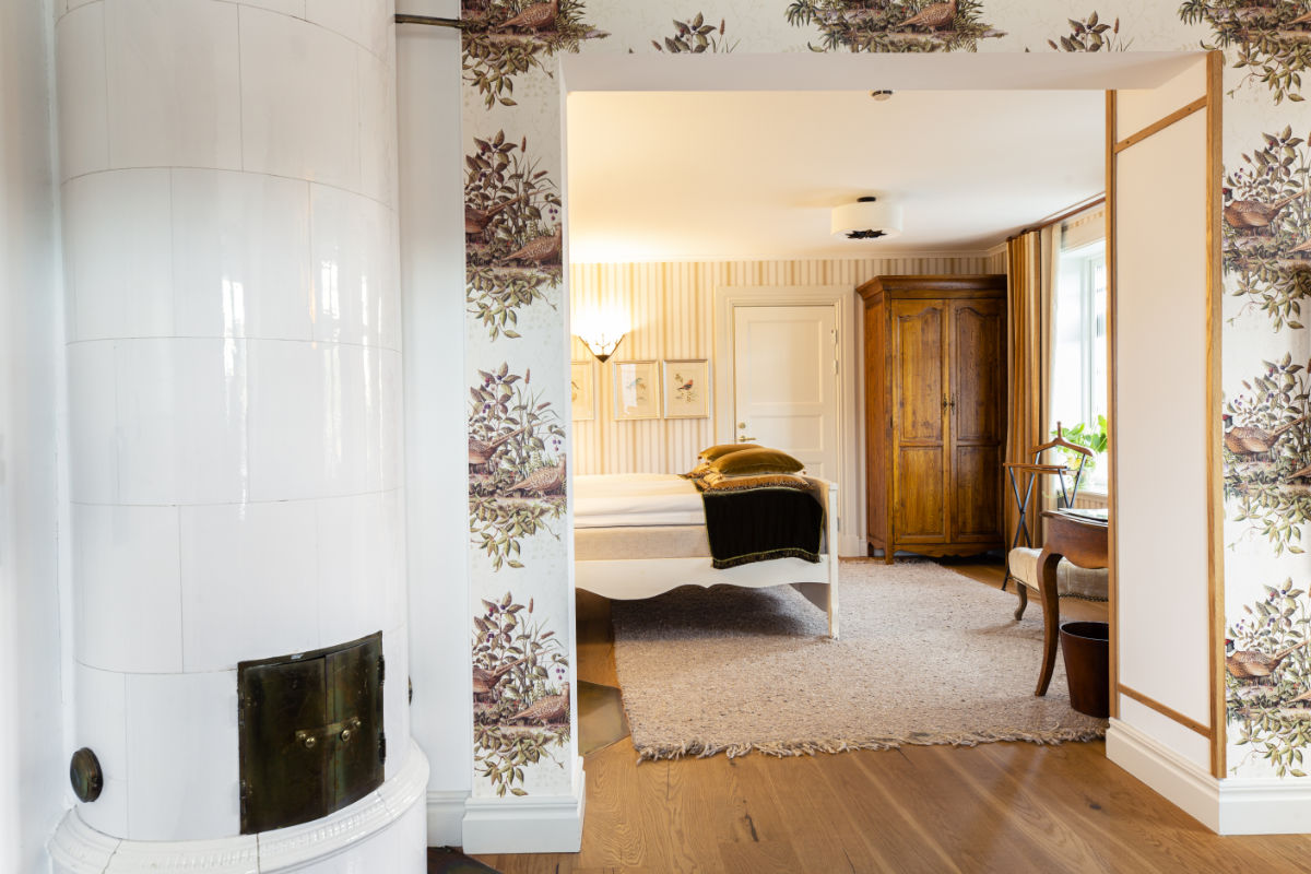 Ein Zimmer im Eriksberg Hotel verfügt über einen weißen Kachelofen im Vordergrund, Blumentapeten, einen Holzschrank, ein Bett mit weißer Bettwäsche und ein großes Fenster, das Tageslicht hereinlässt.
