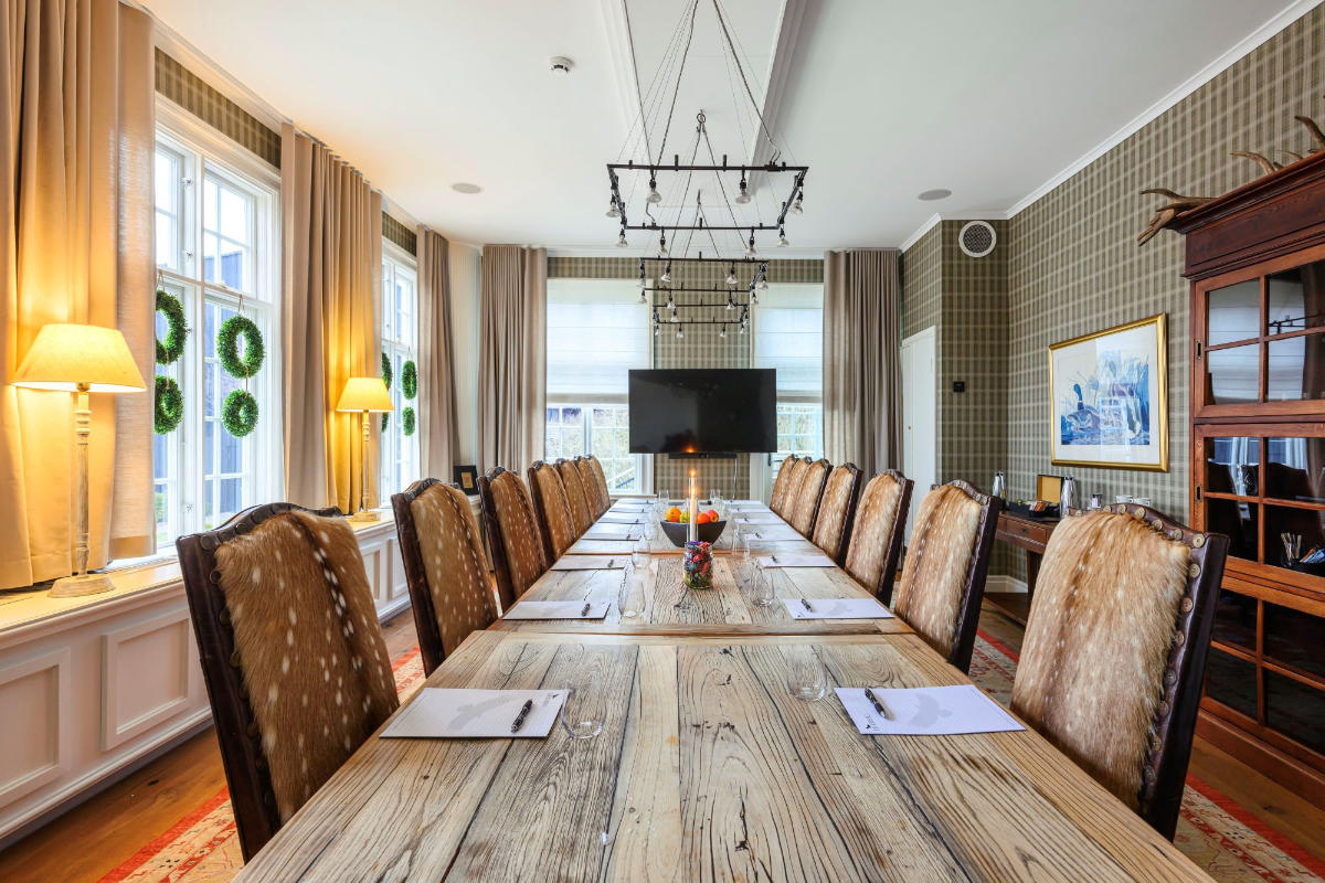 Ein Konferenzraum im Eriksberg Hotel ist mit einem langen Holztisch, Stühlen mit hoher Lehne, einem Fernsehbildschirm am Ende und Fenstern mit Kranzdekoration ausgestattet, die natürliches Licht hereinlassen.