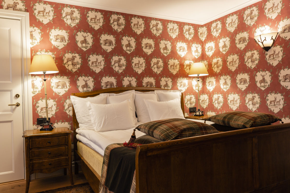 Ein gemütliches Schlafzimmer im Eriksberg Hotel ist mit einem Holzbett, weißen Kissen und karierten Decken ausgestattet. Gemusterte Tapeten mit Vintage-Motiven und zwei Nachttische mit Lampen sorgen für ein warmes Ambiente.