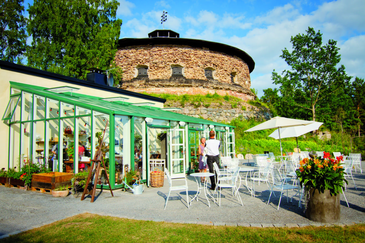 Caffè all'aperto con tavoli e sedie bianche accanto a una serra, di fronte a una fortezza rotonda in pietra circondata da alberi sotto un cielo azzurro.