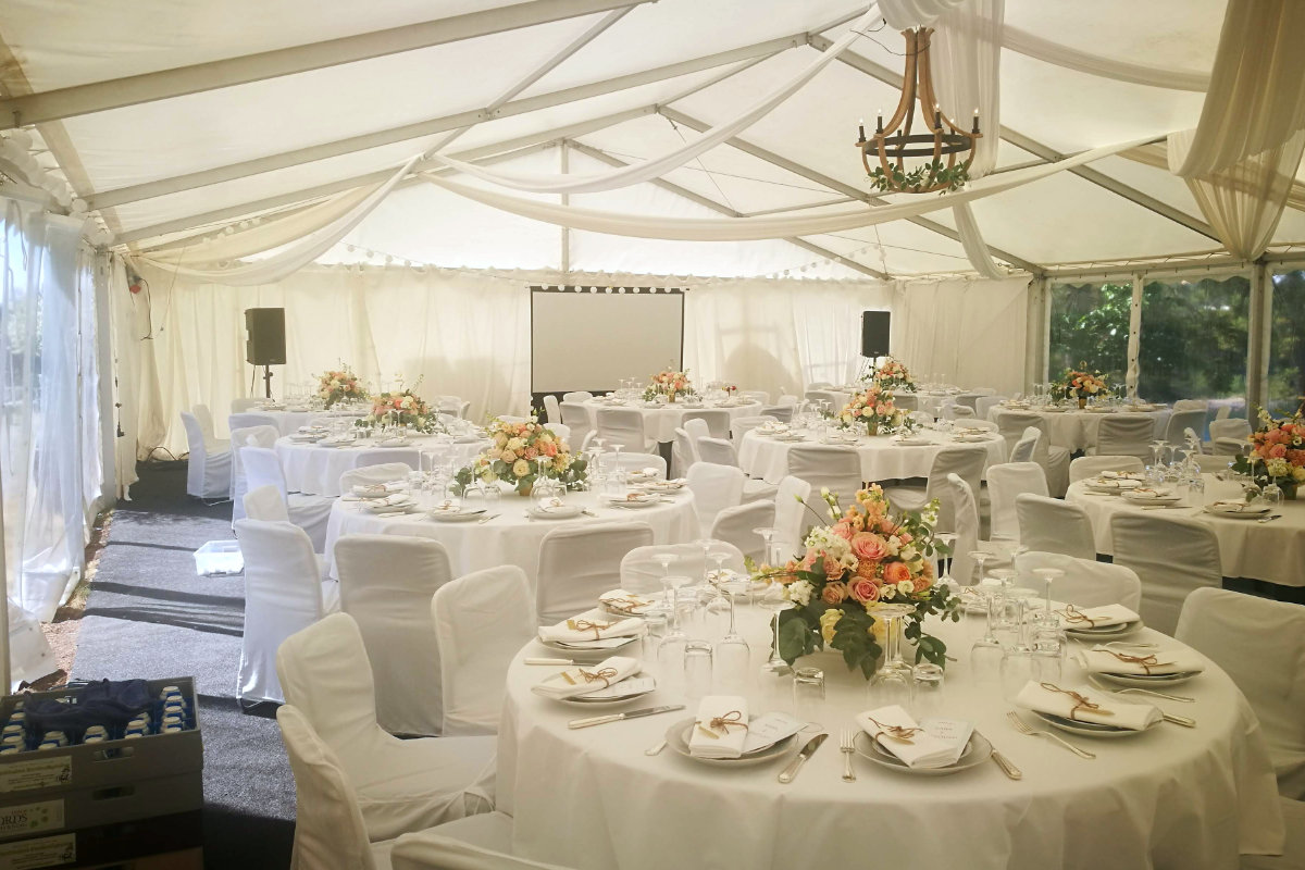 Una tenda bianca per eventi con tavoli rotondi ricoperti di stoffe bianche, apparecchiati per un pasto formale con centrotavola floreali, bicchieri e posate; anche le sedie sono ricoperte di tessuto bianco.