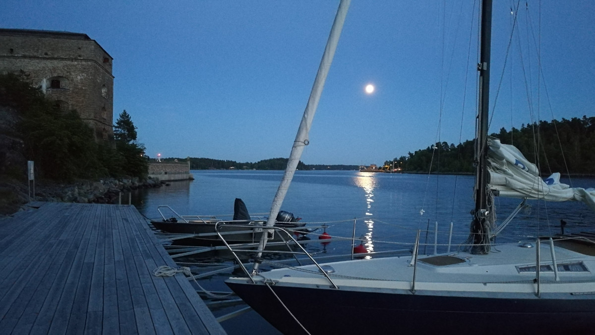 Una barca a vela e un piccolo motoscafo sono attraccati a un molo di legno al tramonto, con l'acqua calma, il cielo illuminato dalla luna e un edificio storico in pietra sulla sinistra.