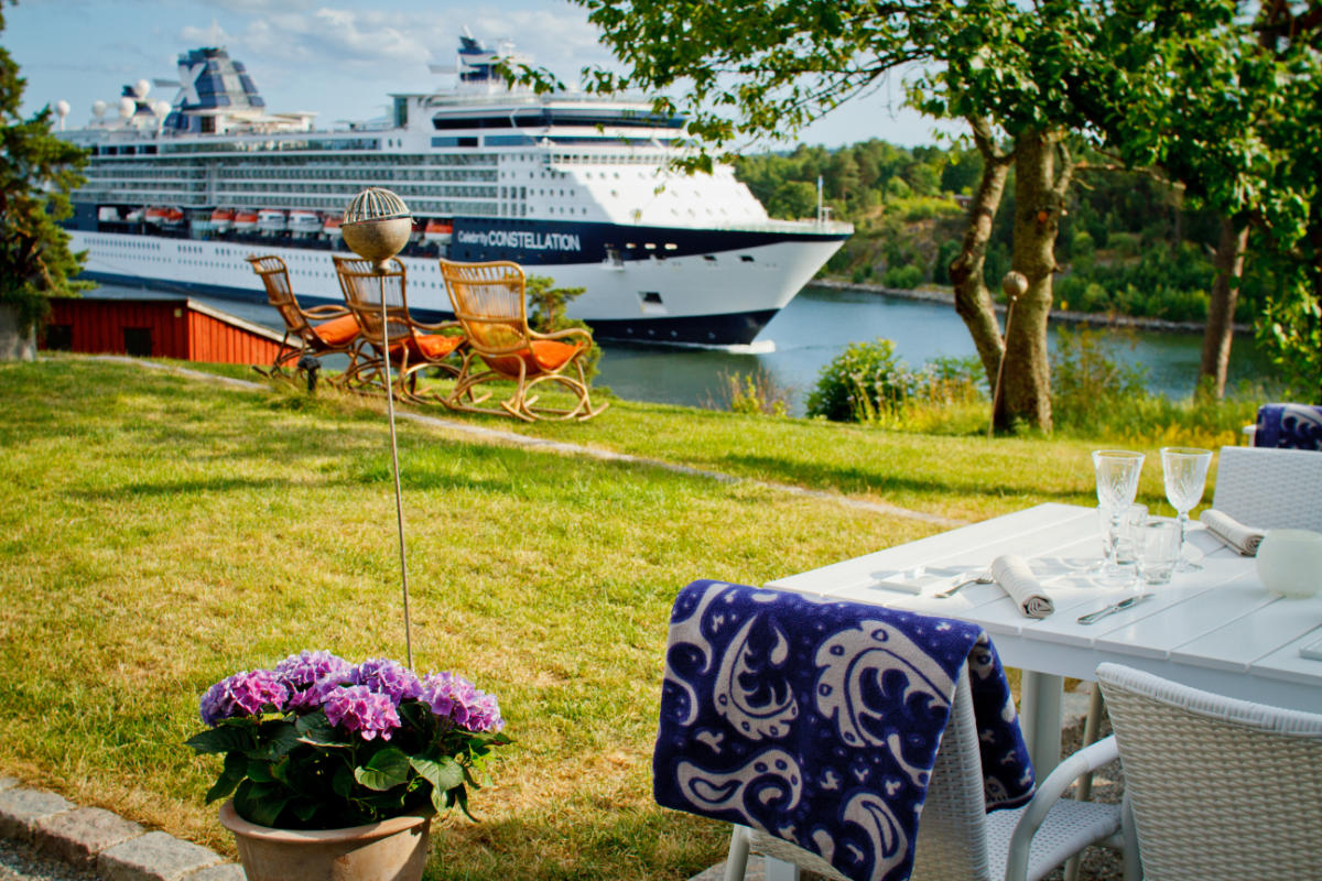 A large cruise ship sails past a grassy yard with a set dining table, wicker chairs, and a potted purple flower in the foreground.