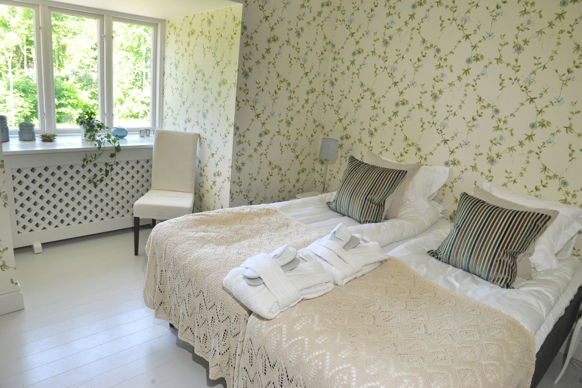 Una luminosa camera da letto con due letti singoli affiancati, ciascuno con copriletto beige, cuscini a righe e asciugamani bianchi piegati; sullo sfondo una finestra e una sedia.