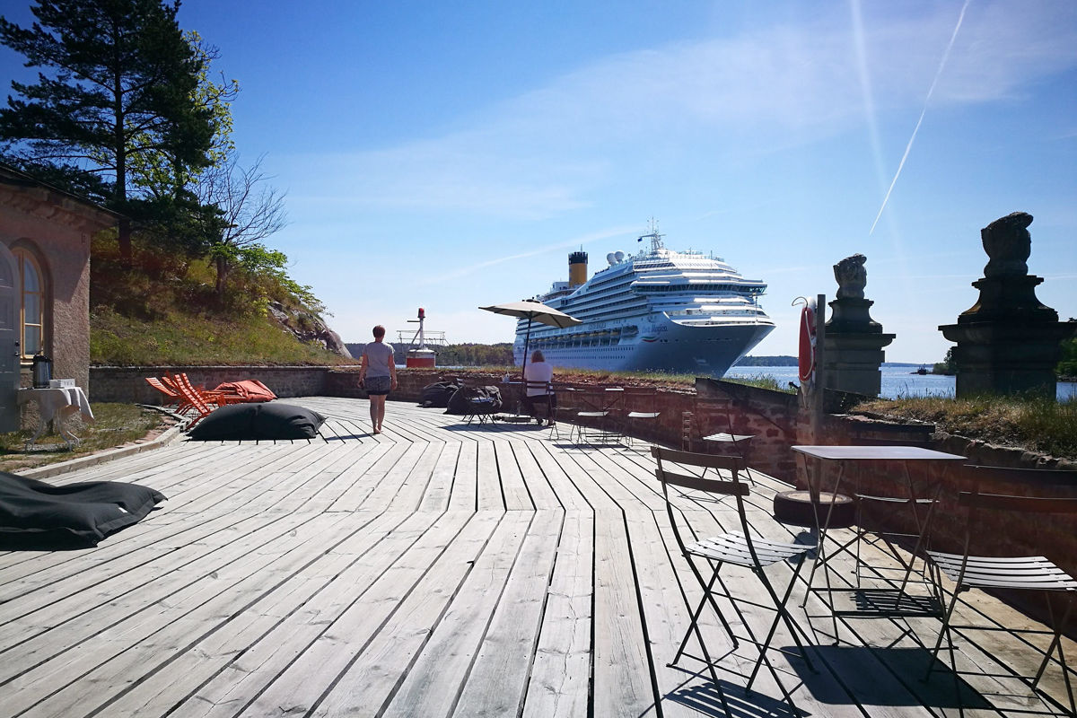Una grande nave da crociera è attraccata vicino a un ponte di legno con sedie, tavoli e salotti presso l'Hotel e la Fortezza di Fredriksborg; due persone camminano sul ponte sotto un cielo azzurro e limpido.