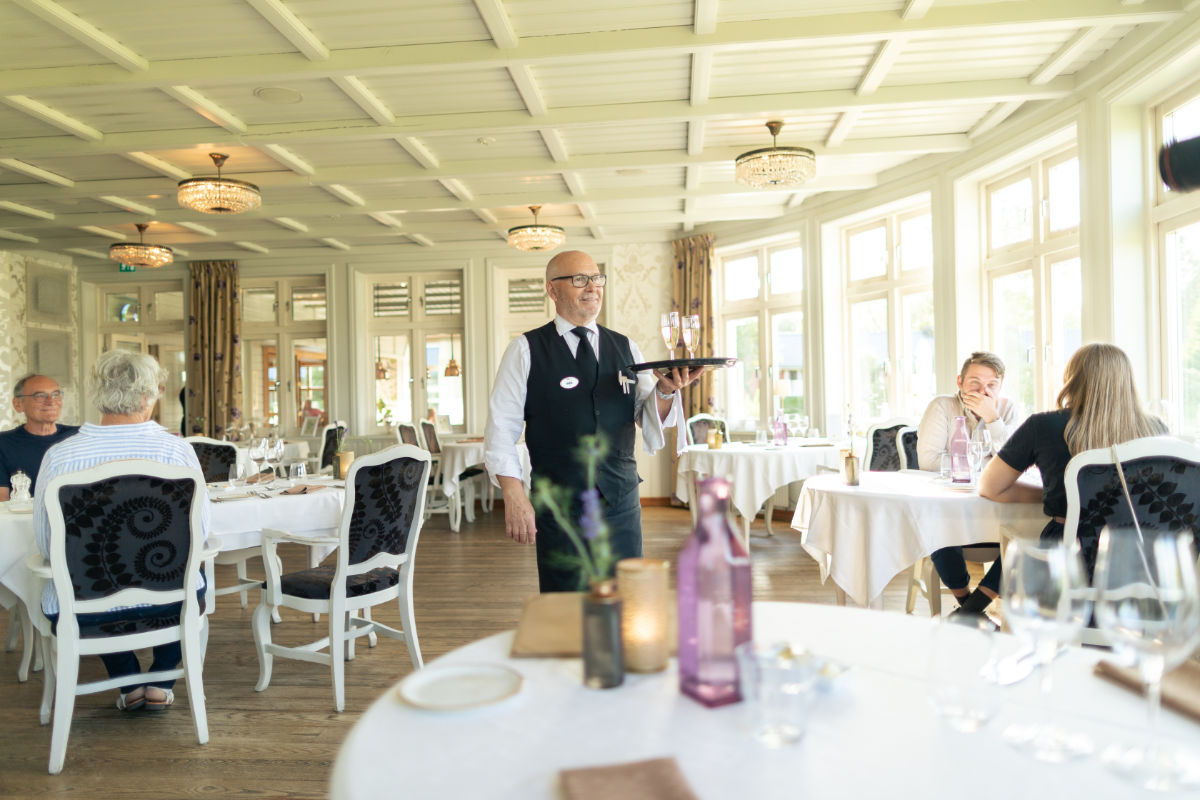A waiter in a black vest carries a tray in a bright, elegant restaurant with several seated patrons and white tablecloths.