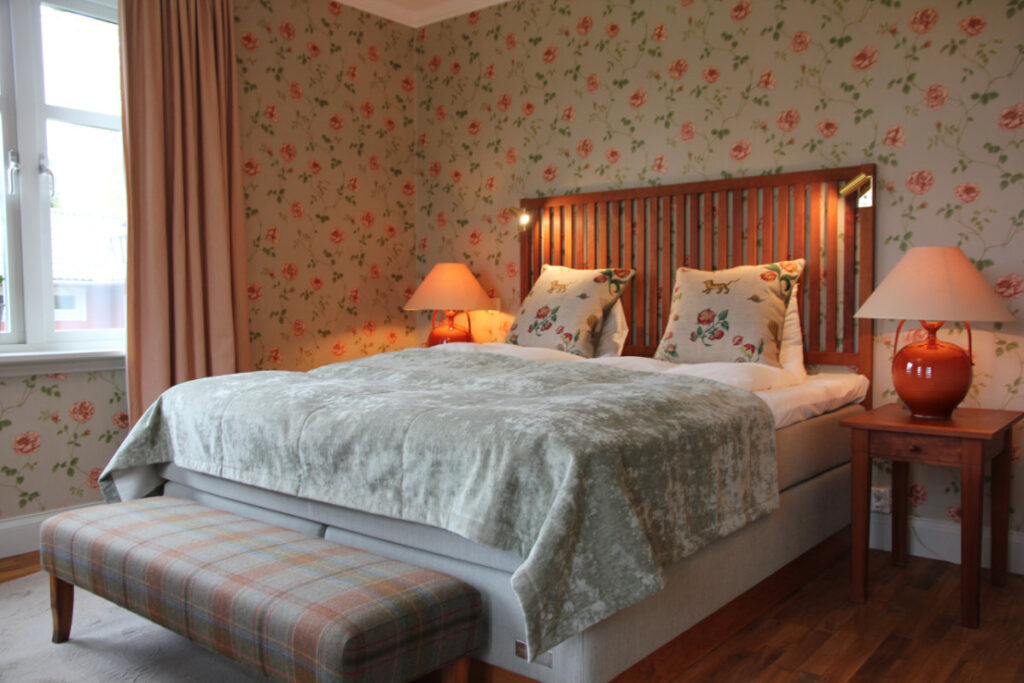 A neatly made bed with floral pillows and a green blanket is flanked by two wooden nightstands with orange lamps in a bedroom with floral wallpaper and a window with beige curtains.