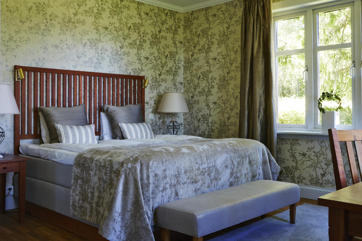 A neatly made double bed with gray bedding and pillows, flanked by two lamps, sits in a room with floral wallpaper, a window with curtains, and a wooden chair.