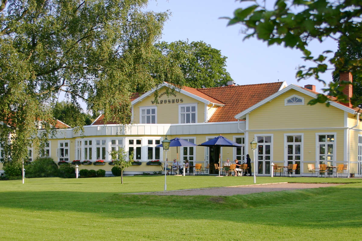 Ein gelbes Holzgebäude mit rotem Ziegeldach, Sitzgelegenheiten im Freien und Sonnenschirmen steht an einem sonnigen Tag auf einer grünen Wiese, umgeben von Bäumen, im Hestraviken Hotel in Schweden.