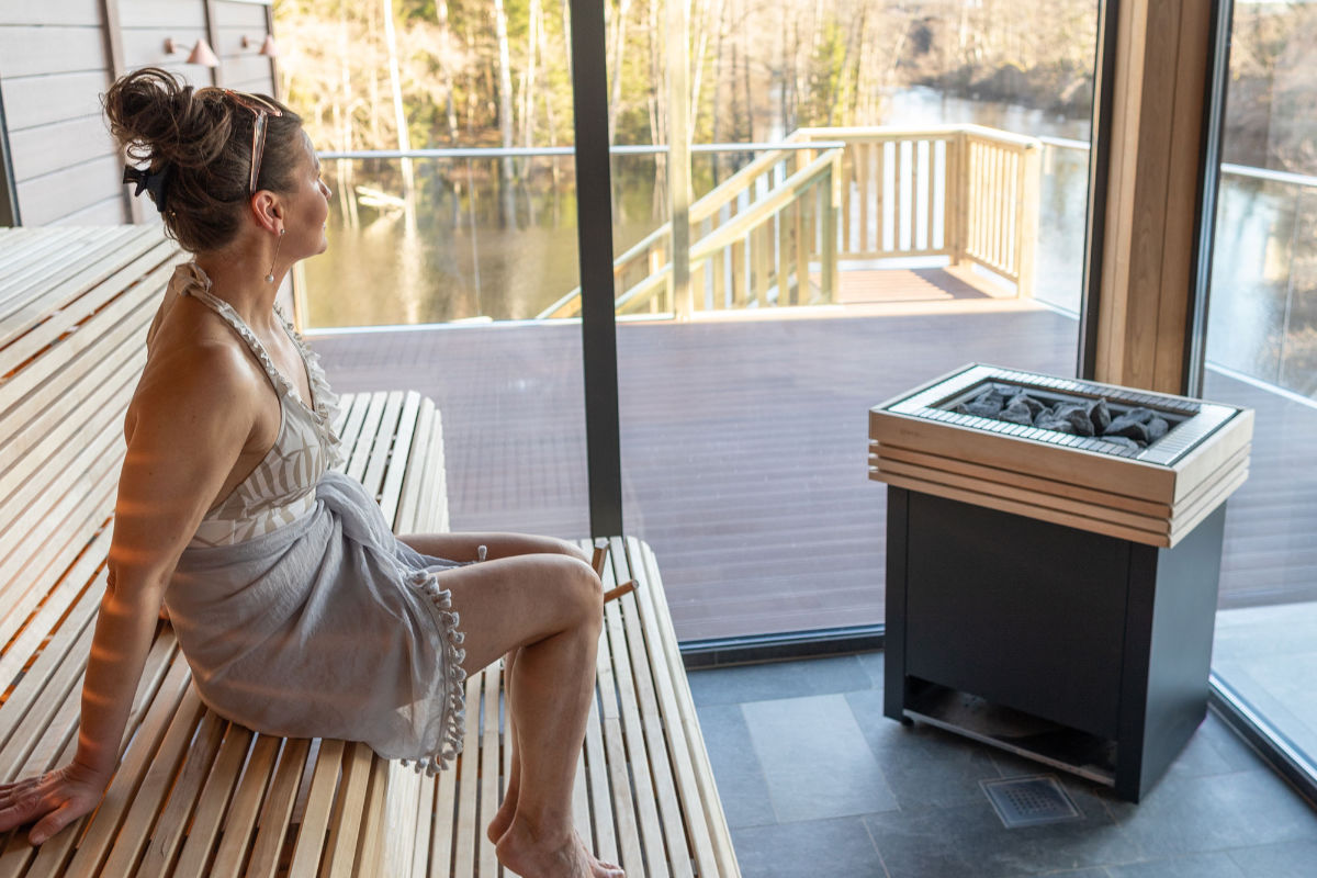 A woman in a towel sits on a wooden bench inside a sauna, looking out through large windows at a deck and trees outside.