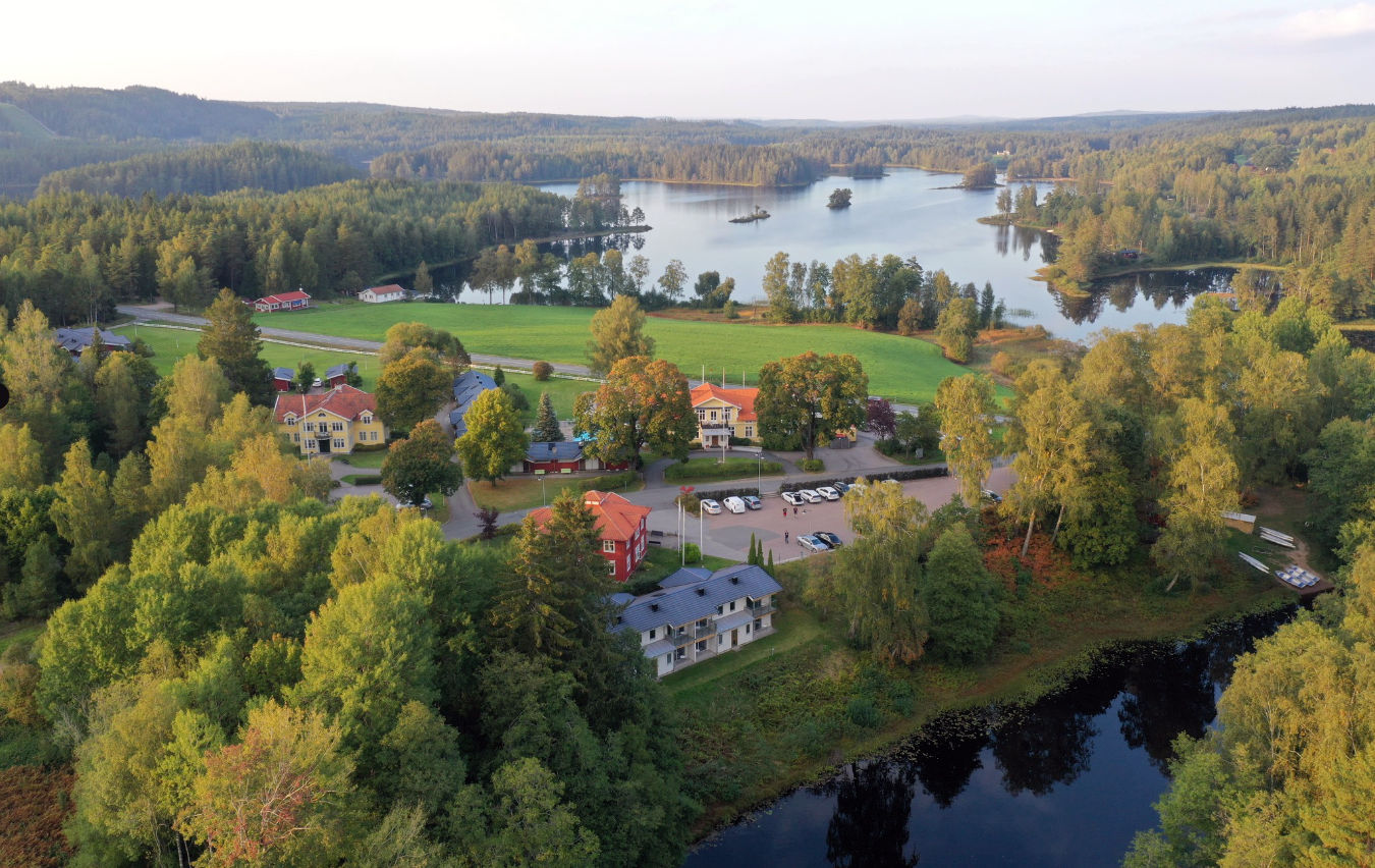 Aerial view of a small lakeside village surrounded by dense forest, grassy fields, and several buildings near the water.