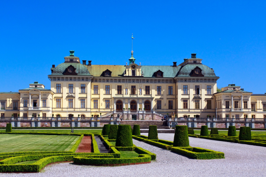 Ein großer, symmetrischer Palast mit gelber Fassade und grünen Kuppeln, umgeben von gepflegten Gärten unter einem klaren blauen Himmel.