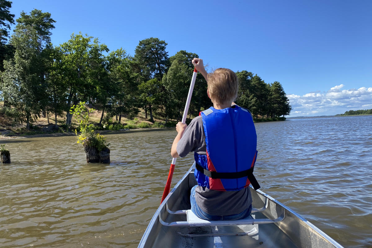 Una persona che indossa un giubbotto di salvataggio blu pagaia una canoa su un lago vicino alla riva alberata del castello di Sundbyholm, sotto un cielo limpido e soleggiato.