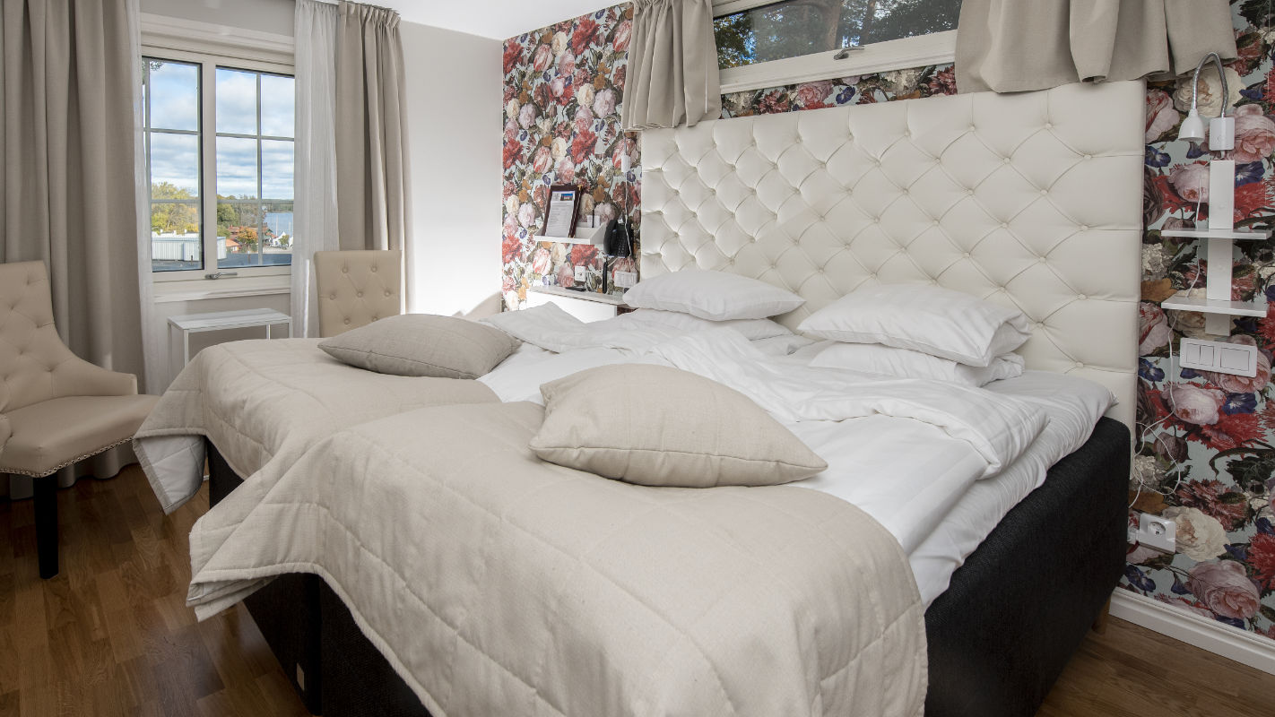 Un letto matrimoniale ben fatto con lenzuola e cuscini beige si trova in una camera con carta da parati floreale, pavimento in legno e due sedie vicino alla finestra, che ricorda il fascino classico del Castello di Sundbyholm.
